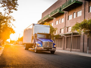 Blue Kenworth box truck driving through an urban commercial district during a local delivery route.