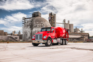 Red Kenworth concrete mixer truck parked outside an industrial concrete production facility with large storage silos and processing equipment in the background.