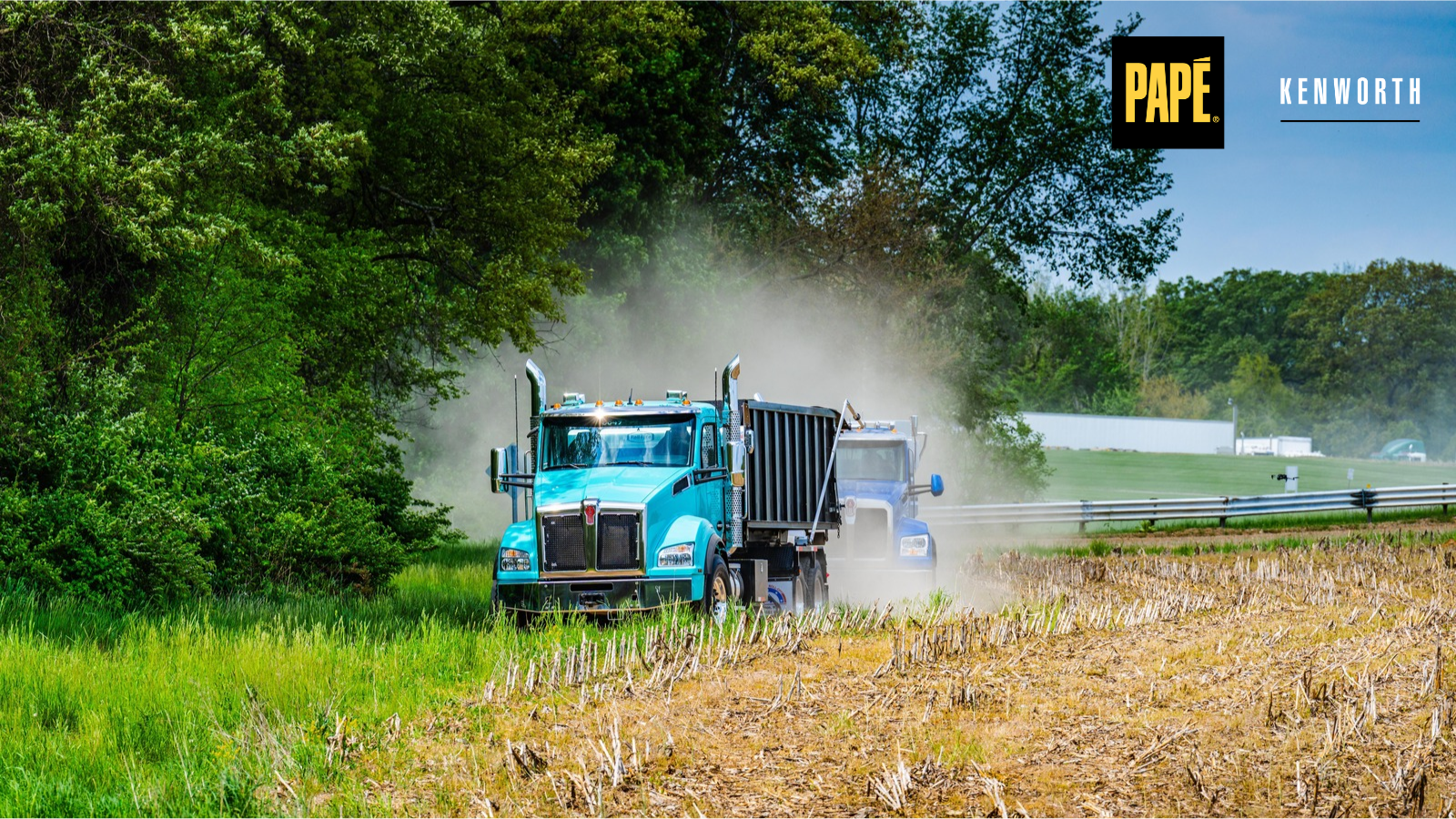 Two Kenworth vocational dump trucks driving along a dirt path beside a field, demonstrating heavy-duty performance in off-road conditions.