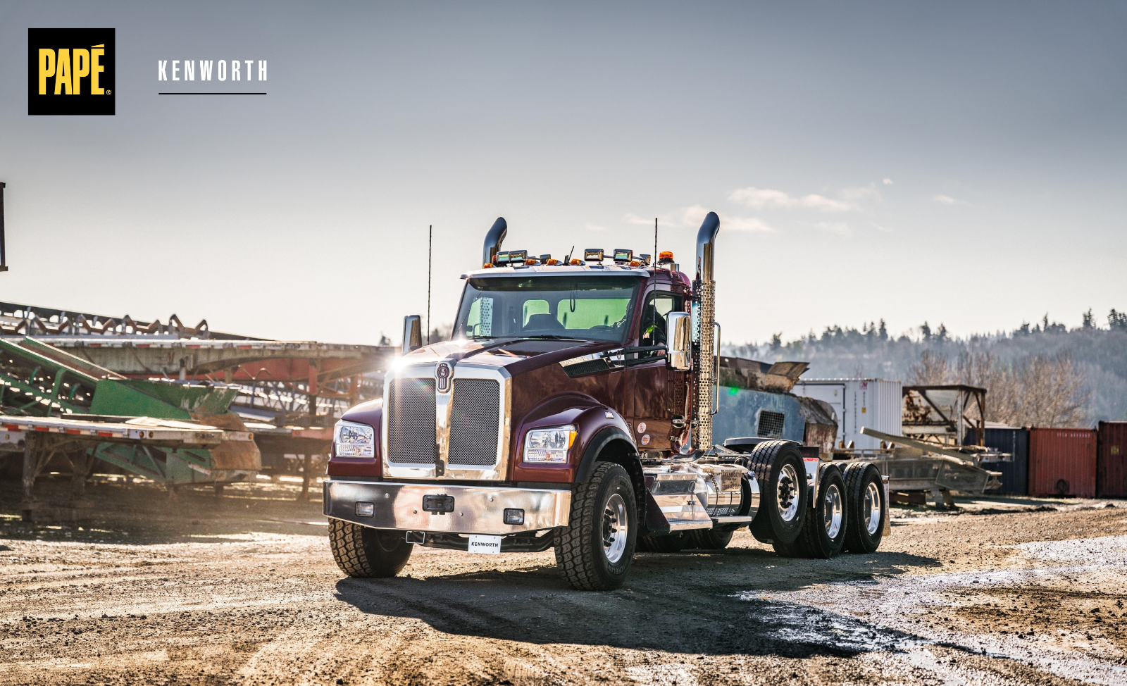 Maroon Kenworth vocational semi-truck with a tandem rear axle parked at an industrial worksite surrounded by heavy equipment.